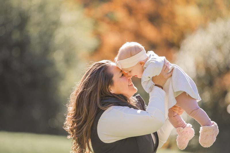 A mother lifts her smiling baby girl toward her face in a sunny autumn field