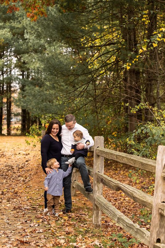 A family gathers along a split-rail fence on a leaf-covered forest path
