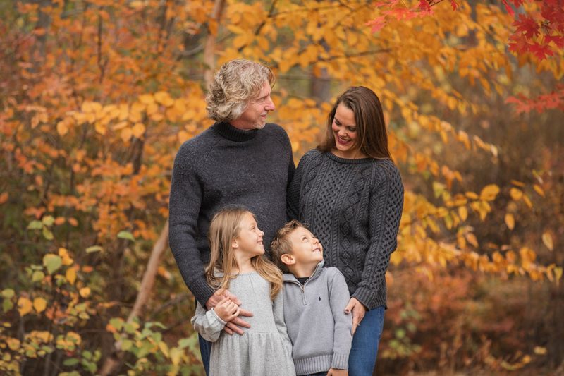 Two children look up at their parents in matching gray sweaters against orange fall foliage