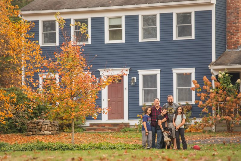 A large family stands on the lawn in front of their blue colonial house in autumn