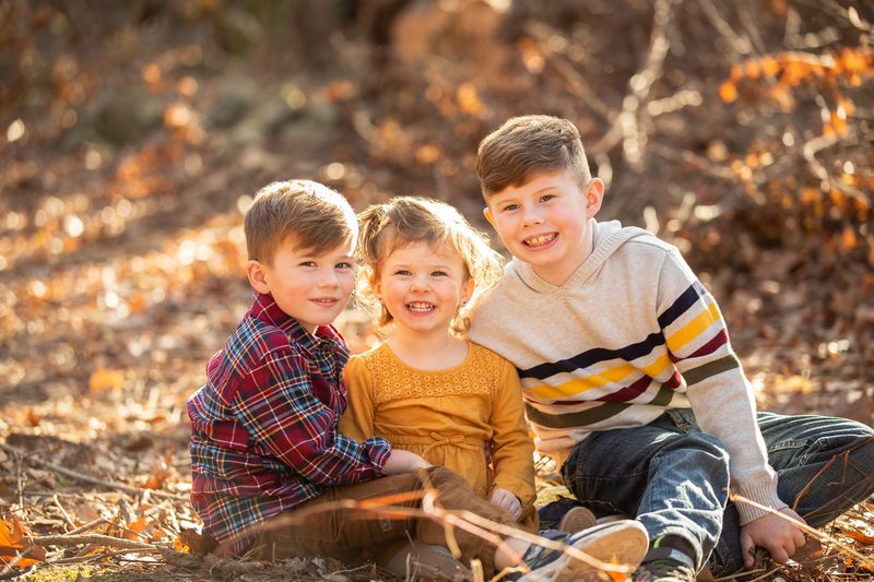 Three young siblings sit grinning together in fallen leaves