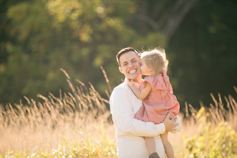 A little girl kisses her laughing dad's cheek in a sunlit golden field