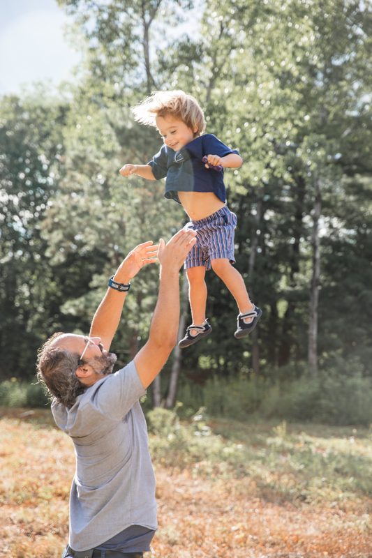 A dad tosses his laughing toddler high into the air in a sunny field