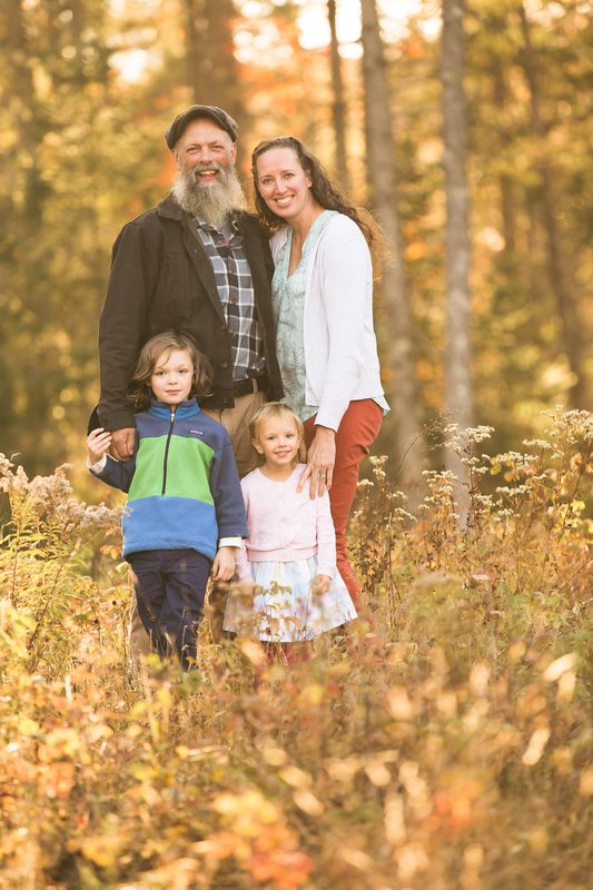 A family of four stands in golden autumn grass at the edge of the woods