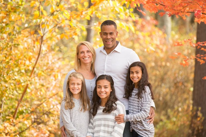 A family of five smiles together in front of bright yellow and red fall trees