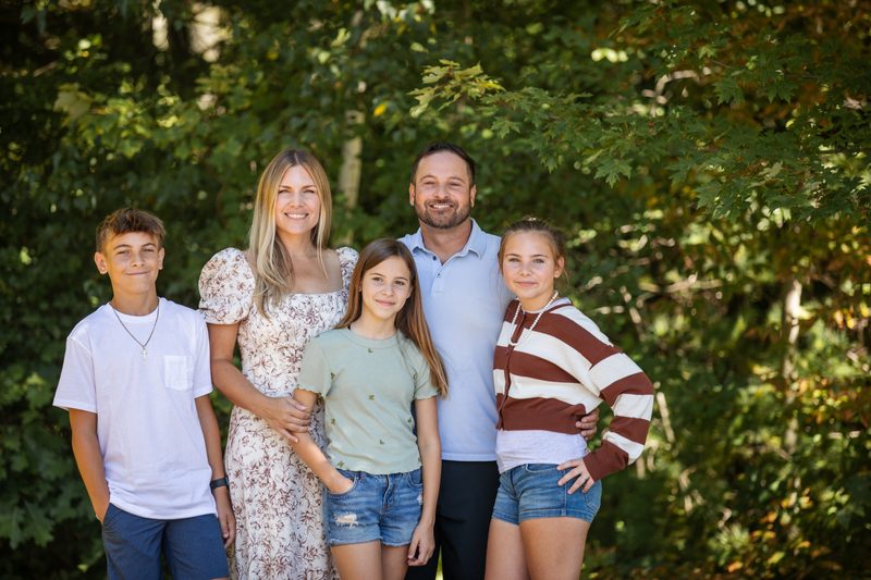 A family of five with three teenagers smiles together in front of leafy summer trees
