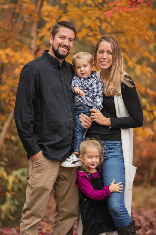 A family of four stands in golden fall woods, dad holding their little boy