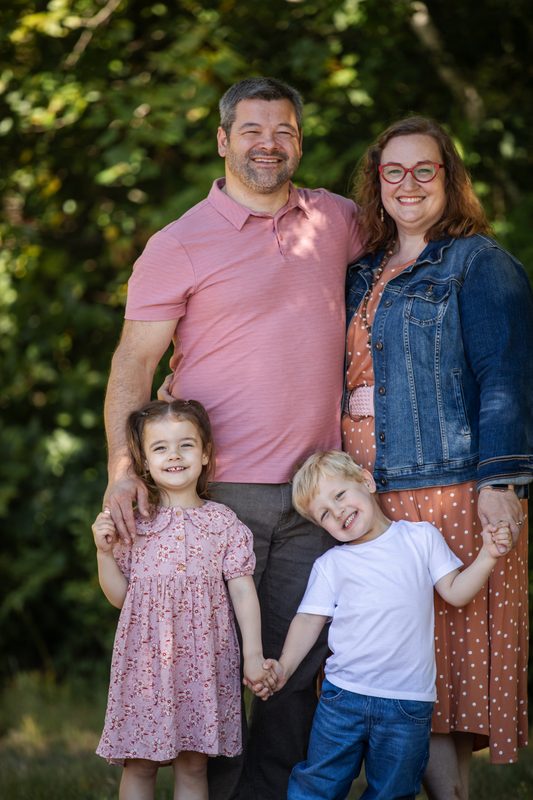 Parents and their two young kids hold hands and grin in front of summer greenery