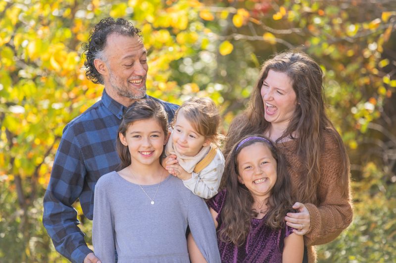 Parents laugh with their three daughters in front of bright golden fall leaves