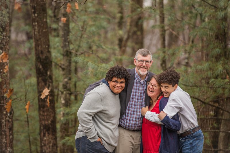 A family of four laughs in a big group hug among tall pine trees