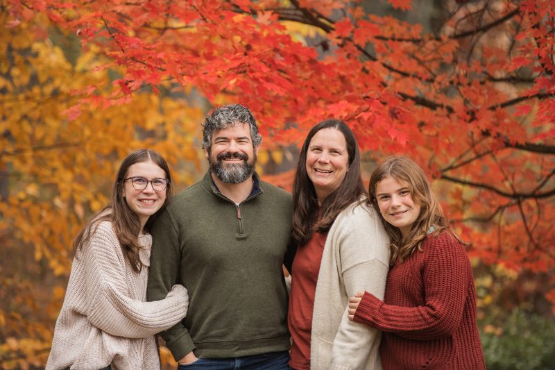 A family of four gathers under a bright red maple tree