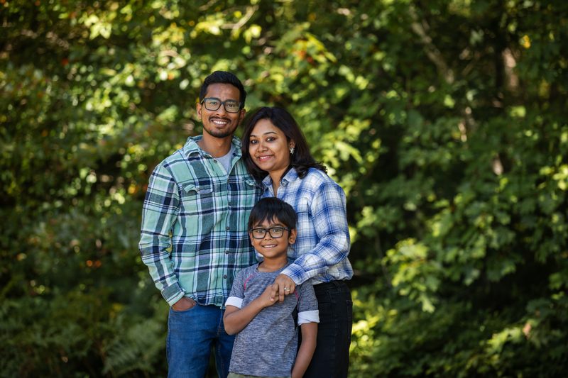 Parents in plaid shirts hug their smiling son in front of green summer trees