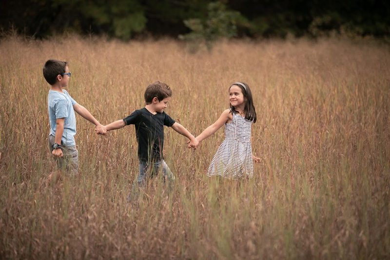Three kids hold hands and walk through a tall golden-grass meadow