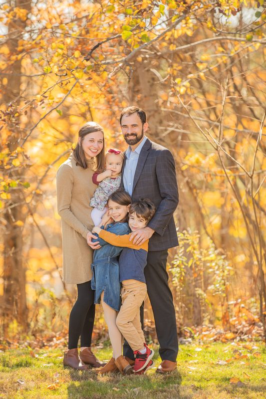 Two children hug their parents in front of golden fall trees