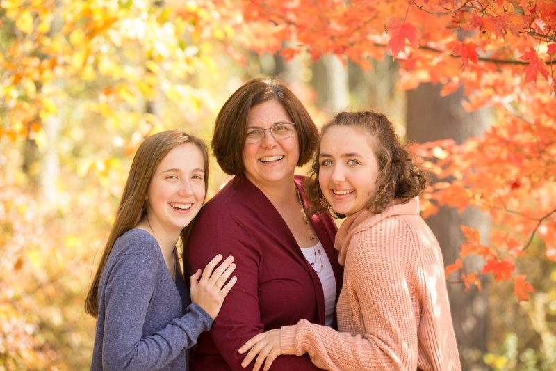 A mother hugs her two daughters under orange and red fall leaves