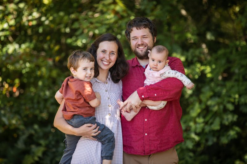 Mom holds her toddler and dad holds their baby girl, all smiling against green trees