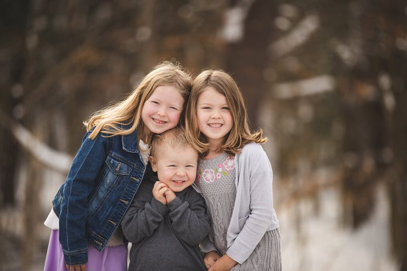 Two sisters and their little brother cuddle close in snowy woods