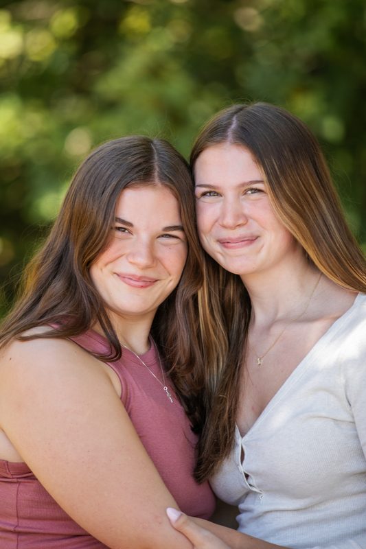Two teenage sisters smile cheek to cheek in a green park