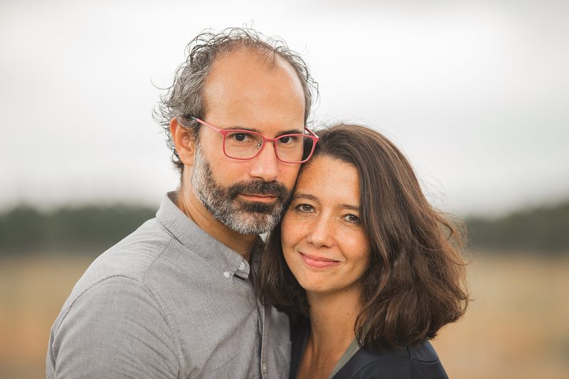 A smiling couple cheek to cheek, him in pink glasses