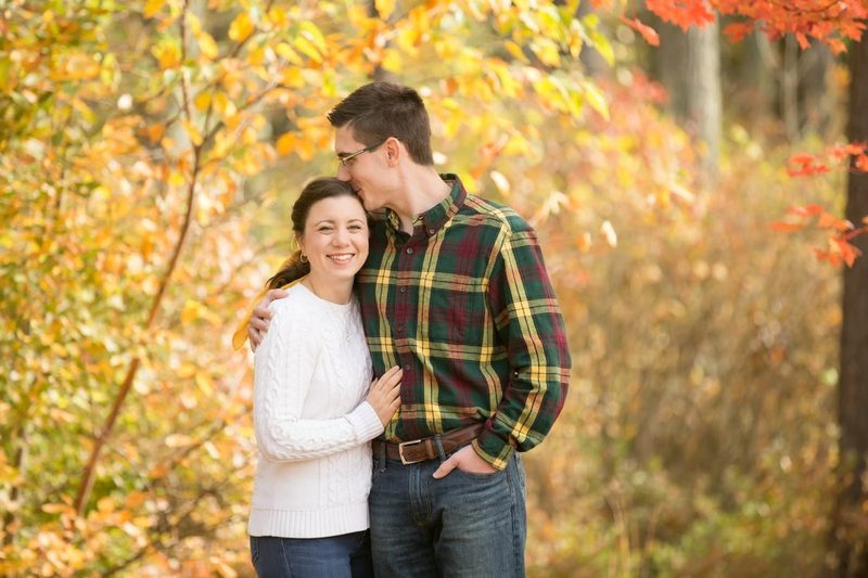 A man kisses his partner's forehead among yellow and red fall leaves