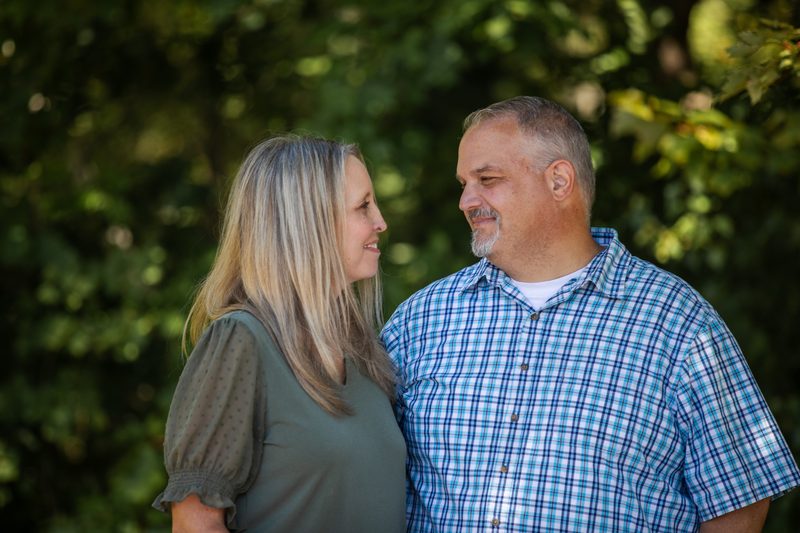 A couple smiles at each other in front of sunlit summer trees