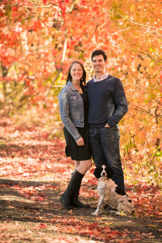 A smiling couple stands on a leaf-covered path under red fall trees, their spaniel sitting at their feet
