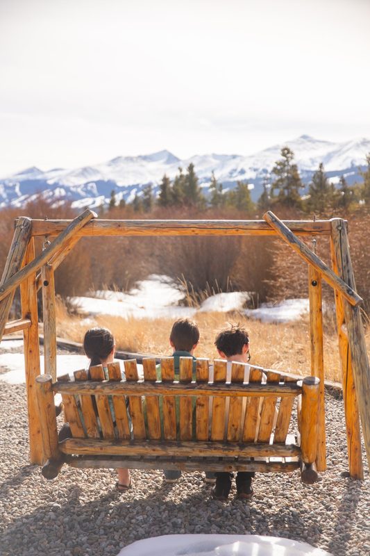 Three kids sit on a wooden swing facing snowy mountains