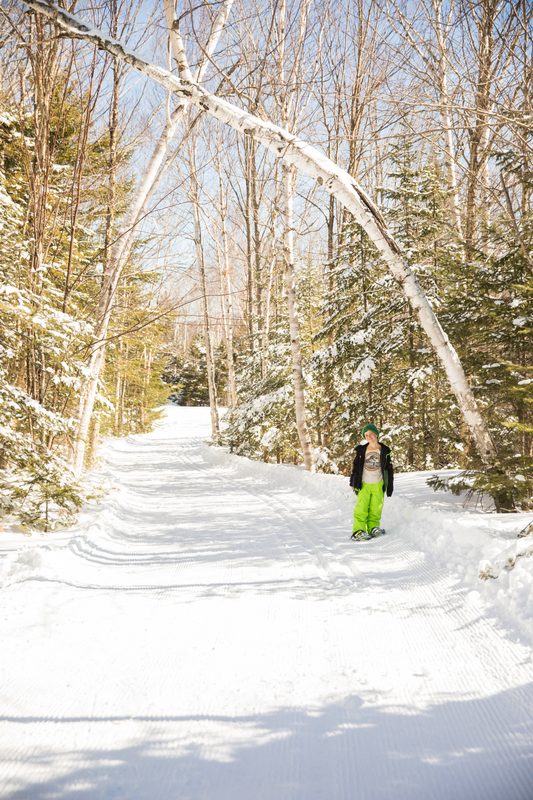 A child in bright green snow pants walks a snowy trail through birch trees